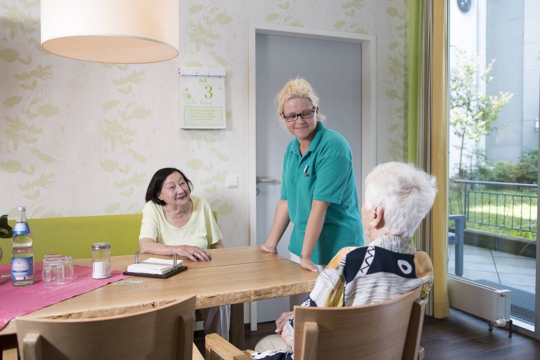 Eine Pflegekraft unterhält sich freundlich mit zwei älteren Damen an einem Tisch.