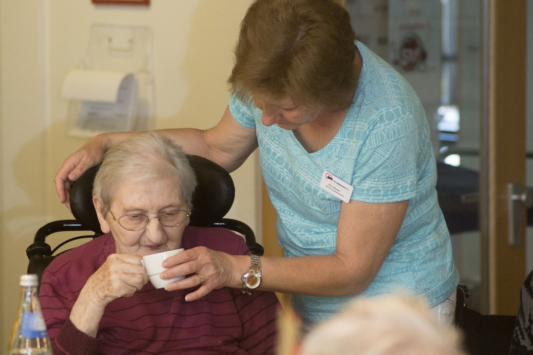 Eine Pflegekraft hilft einer Seniorin beim Trinken aus einer Tasse.