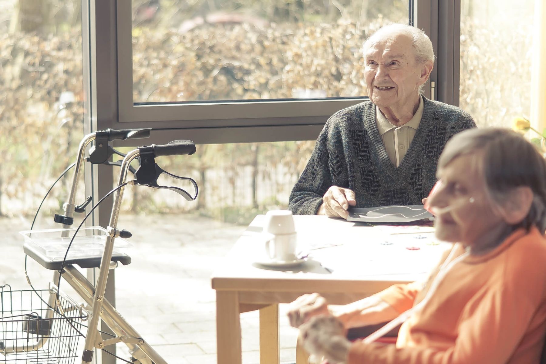 Zwei Senioren sitzen am Fensterplatz, einer nutzt einen Rollator.