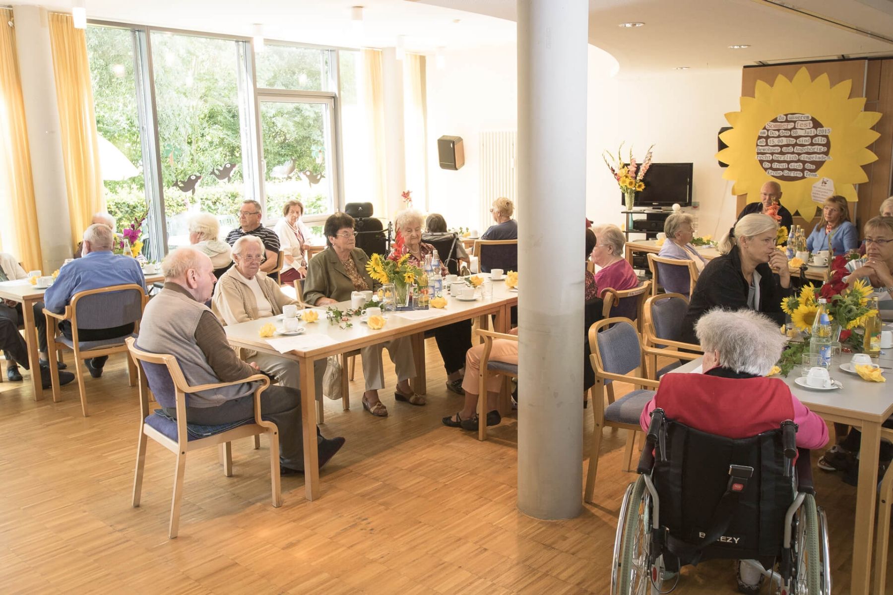 Viele Senioren sitzen in einem Gemeinschaftsraum bei Kaffee und Blumen.