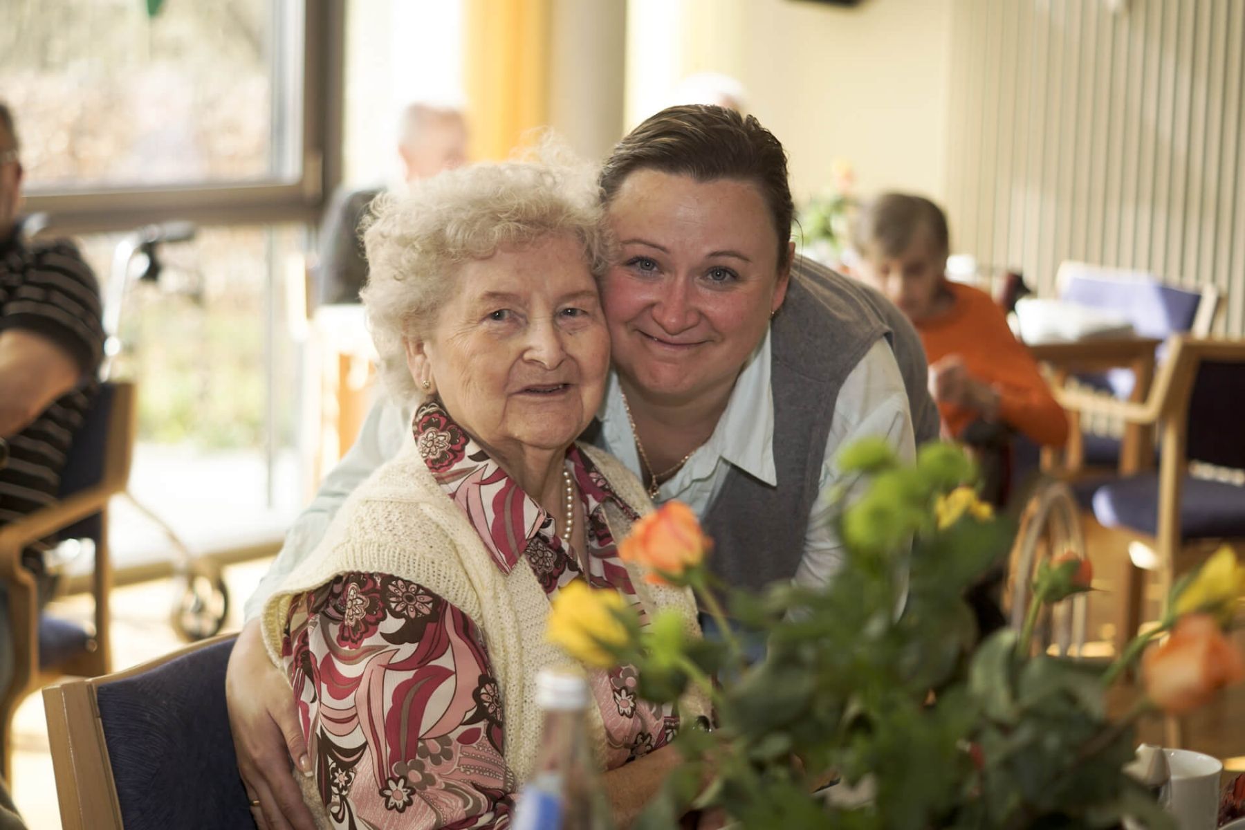Eine ältere Frau sitzt lächelnd mit einer Pflegerin Arm in Arm in einem Gemeinschaftsraum.