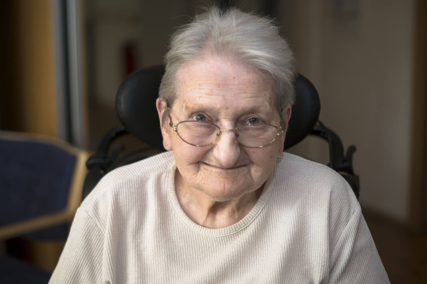 Eine Seniorin mit Brille sitzt in einem Rollstuhl und lächelt direkt in die Kamera.