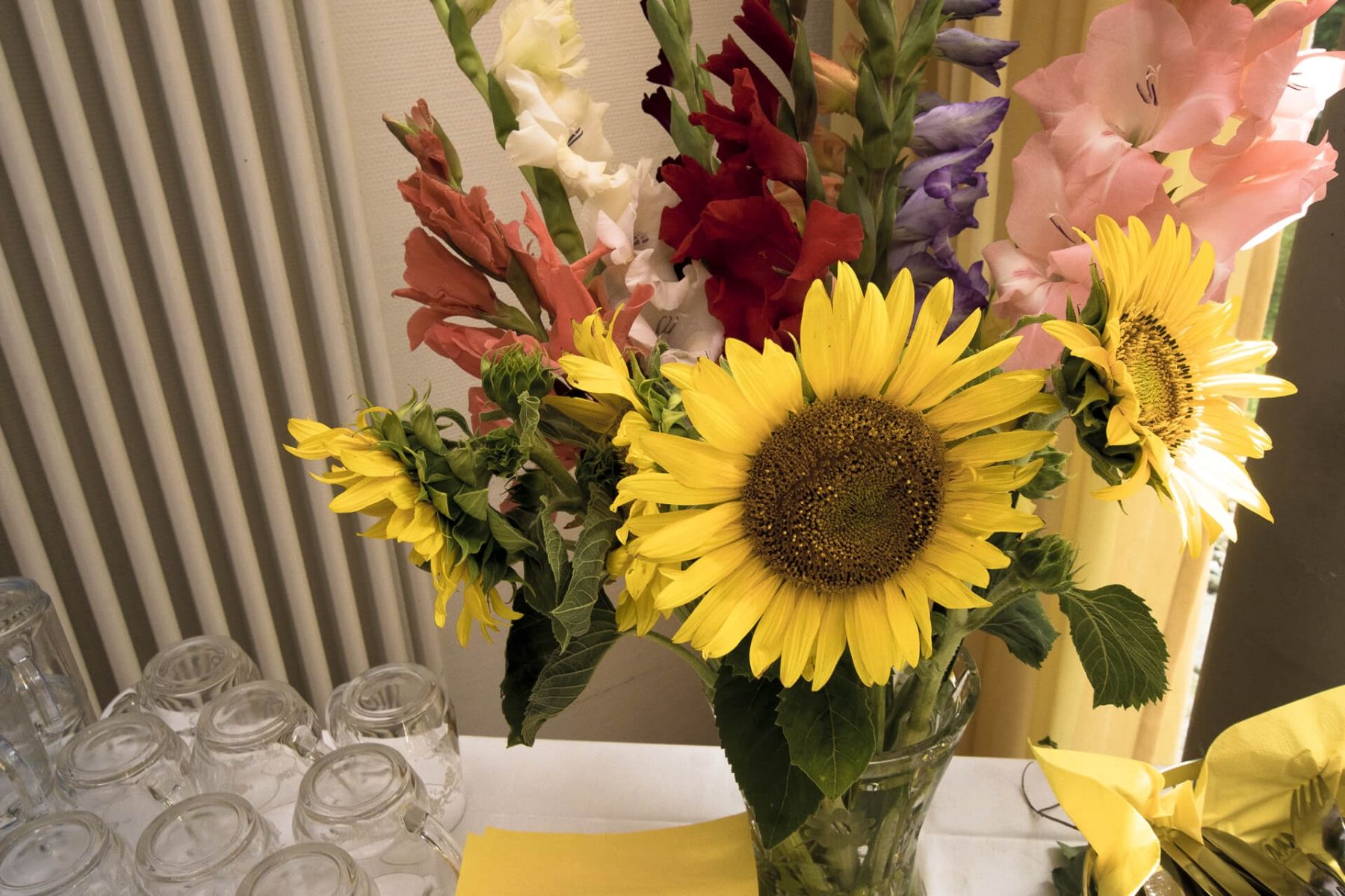 Ein bunter Blumenstrauß mit Sonnenblumen und Gladiolen steht auf einem Tisch neben mehreren Gläsern.