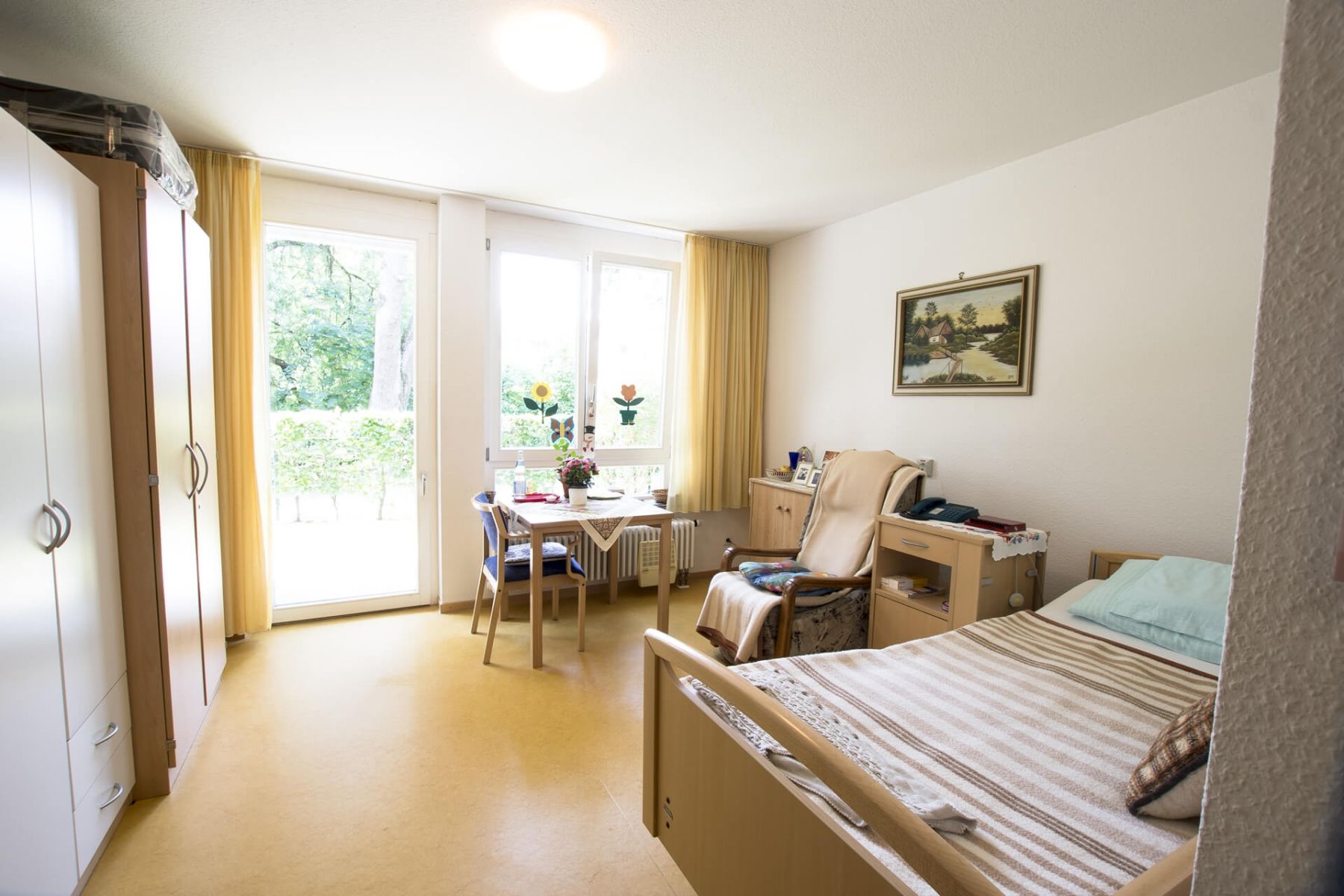 Helles, gemütlich eingerichtetes Einzelzimmer mit Tisch, Bett und großen Fenstern.