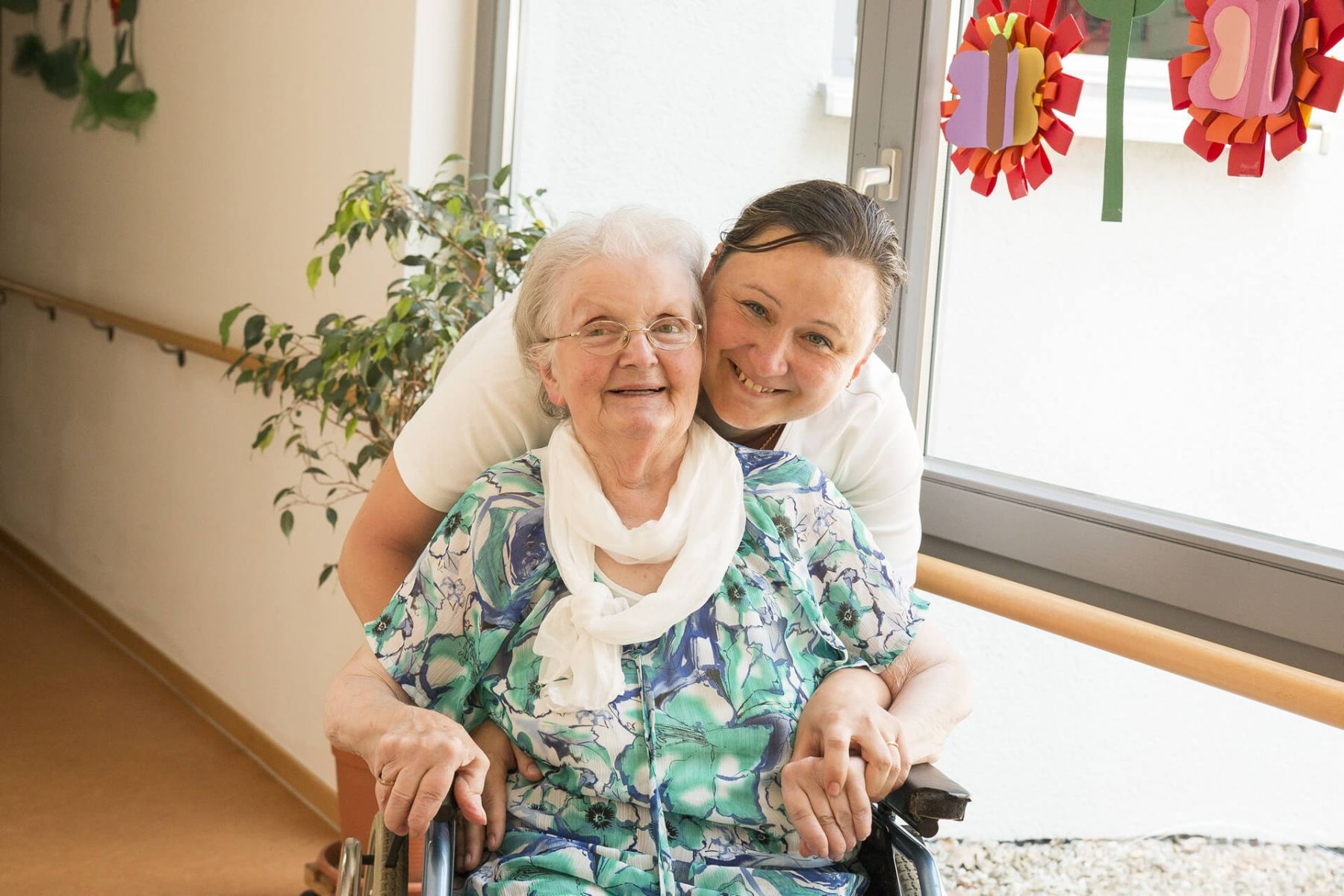 Glückliche Seniorin im Rollstuhl mit freundlicher Pflegekraft im Flur des Pflegeheims.