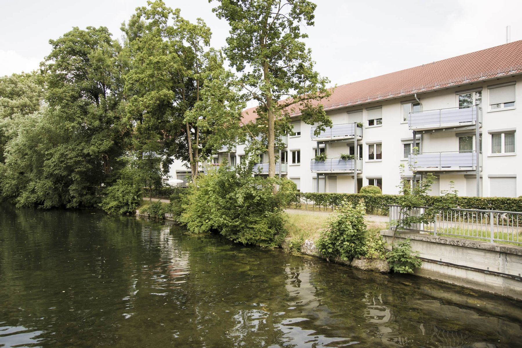 Ein modernes Wohngebäude liegt idyllisch an einem Flussufer, umgeben von Bäumen.